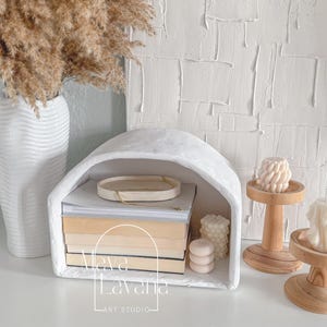 May include: White arched shelf with a stack of books and candles. The shelf is made of plaster and has a rustic look. The shelf is labeled "Viva La Vania Art Studio".
