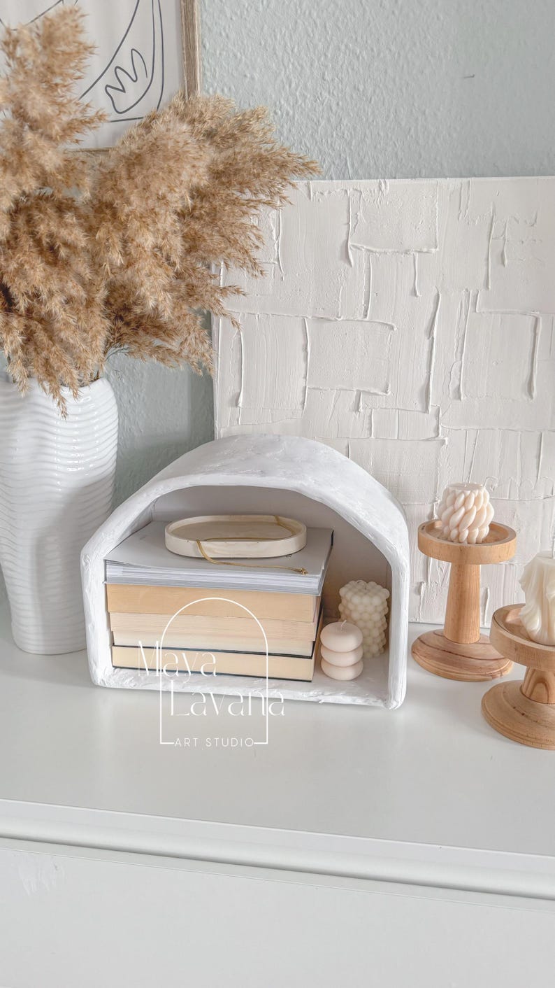May include: White arched shelf with a stack of books and candles. The shelf is made of plaster and has a rustic look. The shelf is labeled "Viva La Vania Art Studio".