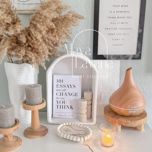 May include: A white shelf with a white archway, a book titled "101 Essays that will Change the way You Think" by Brianna Wiest, three wooden candle holders, a white ceramic tray with a gold chain, a glass candle holder, and a wooden essential oil diffuser.