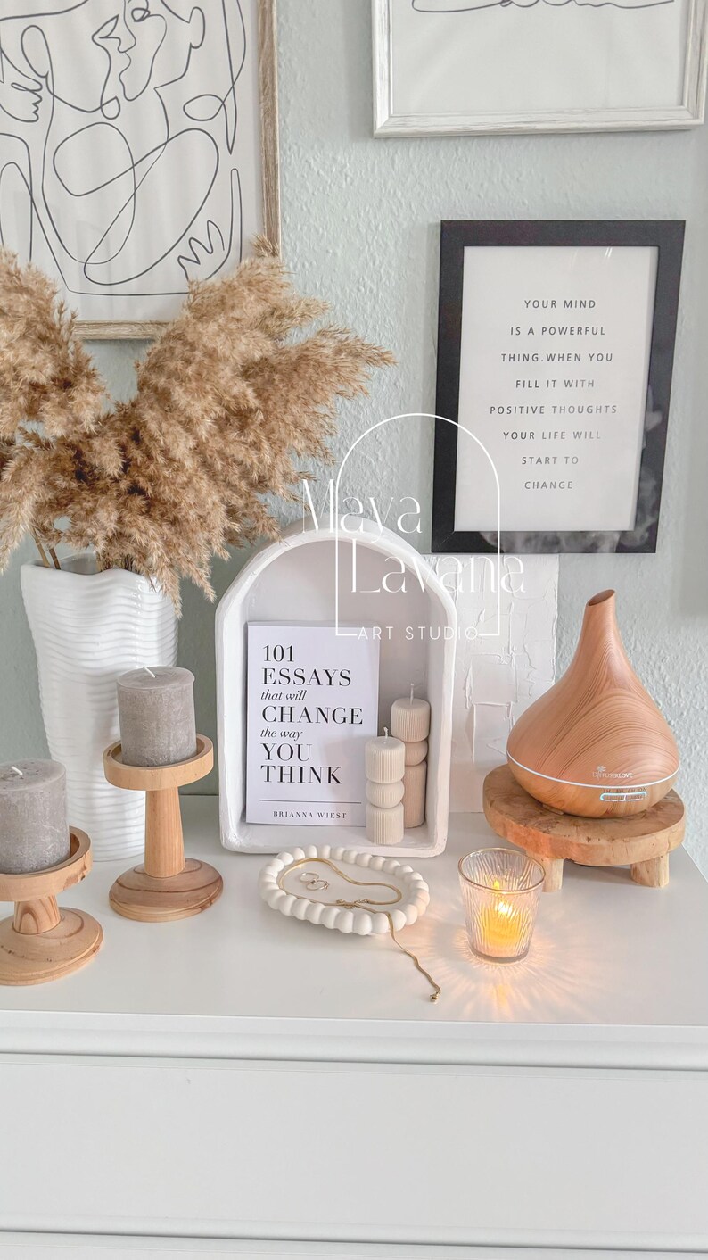 May include: A white shelf with a white archway, a book titled "101 Essays that will Change the way You Think" by Brianna Wiest, three wooden candle holders, a white ceramic tray with a gold chain, a glass candle holder, and a wooden essential oil diffuser.