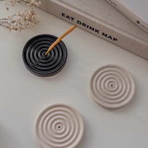 May include: Three round incense holders, two are white and one is black. The black holder has a wooden incense stick in it. The holders have a concentric circle design.