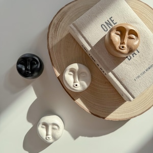 Könnte beinhalten: Drei Keramik-Räucherstäbchenhalter in Form von Gesichtern. Die Halter sind in Schwarz, Weiß und Braun. Die Halter stehen auf einer Holzoberfläche mit einem Buch, auf dem "One Line a Day, A Five Year Memory" steht.