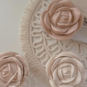 May include: Three large, beige, fabric rose flower decorations on a white, crocheted, round mat with fringe. The flowers are arranged in a semi-circle. The text "Aesthetic Sustainable Home Decor" is in the lower left corner of the image.