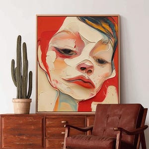 Puede incluir: Pintura abstracta de un rostro con tonos rojos, blancos y naranjas, enmarcada en madera clara. Una silla moderna de mediados de siglo marrón y un mueble de madera con un cactus en maceta están en primer plano.