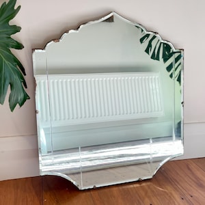 May include: A wall mirror with a scalloped top edge and a multi-paneled glass design. The mirror features visible signs of age and wear along the edges of the frame, with a clear reflective surface showing reflections of a radiator and plant leaves.