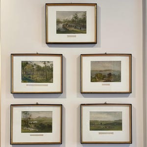 May include: Five framed landscape prints with black and gold frames are displayed on a white wall. The prints depict scenes of nature, including trees, water, and fields. A wooden chest with ornate legs sits below the artwork.