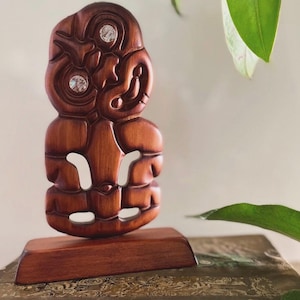 Könnte beinhalten: Eine geschnitzte Hei Tiki-Figur aus Holz mit poliertem braunem Finish und zwei eingelegten, schillernden Muschelaugen. Die Figur zeigt traditionelle stilisierte Gesichtszüge und ein durchbrochenes Körperdesign auf einem rechteckigen Holzsockel.