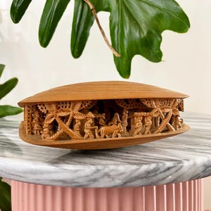 Puede incluir: Una caja de madera tallada con tapa, con intrincados relieves de personas, palmeras y animales. La caja, de forma ovalada, descansa sobre una superficie de mármol. La madera es de color marrón claro cálido.