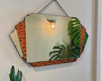 May include: Vintage Art Deco style mirror with a geometric shape. The mirror has a central rectangular section and side panels with a textured, orange-brown border. It hangs from a chain and features a small light fixture.