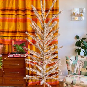 Può includere: Un albero di Natale artificiale, alto e sottile, con rami bianchi e piumosi. L'albero è su un supporto in metallo argentato. Lo sfondo include una tenda a righe arancioni e gialle in stile retrò e un comò in legno.