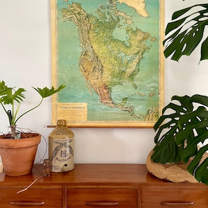 Könnte beinhalten: Eine Vintage-Karte von Nordamerika hängt an der Wand. Die Karte ist in Grün-, Braun- und Gelbtönen gehalten und hat einen Holzrahmen. Unter der Karte steht eine braune Holzkommode mit einem Keramikkrug und einer Topfpflanze.