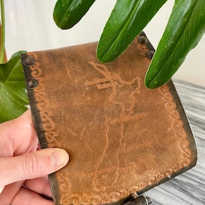 May include: A brown leather wallet with a decorative border and a clasp closure. The wallet has a textured surface and appears to be handmade. The design includes embossed details and rivets along the edges. The wallet is held in front of a green leafy plant.