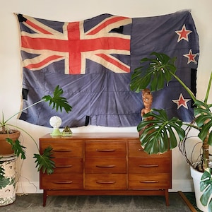 May include: A mid-century wooden dresser with nine drawers, beneath a faded blue flag with a red Union Jack and stars. The room is decorated with potted plants and decorative objects.