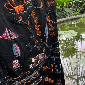 May include: A close-up shot of a black fabric with detailed beadwork. The design incorporates floral and figurative patterns, with orange, pink, and blue beads. The embroidery creates a textured surface, showcasing a variety of colours and shapes.