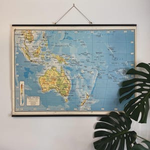 May include: A vintage map of the Indo-Pacific region, featuring Australia, New Zealand, and Southeast Asia. The map is printed on a blue background with green and brown landmasses. The map is framed in a black frame and is hanging on a white wall.