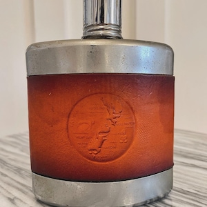 Könnte beinhalten: Eine silberne und braune Leder-Hüftflasche mit einem runden Design, das eine Karte von Neuseeland und den Text "NEW ZEALAND" und "WELLINGTON" zeigt.