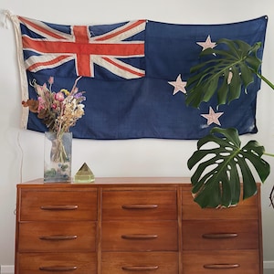 Pode incluir: Uma bandeira vintage da Nova Zelândia com fundo azul, cruz vermelha e estrelas brancas está pendurada acima de uma cômoda de madeira. Um vaso transparente com flores secas e uma pirâmide de vidro verde estão na cômoda. Grandes folhas verdes de Monstera à direita.