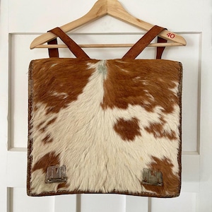 Puede incluir: Una mochila de piel de vaca marrón y blanca con correas de cuero. La mochila tiene una forma rectangular y está hecha de un material suave y natural. Las correas son ajustables y tienen un cierre de hebilla. La mochila es un accesorio único y elegante para el uso diario.