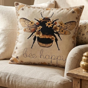 May include: A beige armchair with a decorative pillow featuring a large bee illustration with floral accents and the words "bee happy." Additional pillows and a small side table with books and a vase of flowers complete the cozy scene.