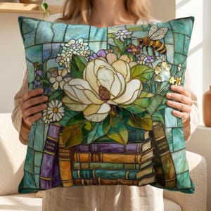 Può includere: Cuscino quadrato decorativo con un design a vetrata raffigurante un grande fiore di magnolia bianca, un'ape e una pila di libri. Il cuscino ha uno sfondo blu e turchese con dettagli floreali e di libri.