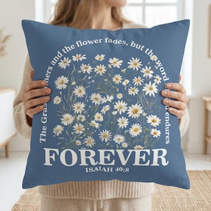 Puede incluir: Un cojín cuadrado azul con un diseño floral de margaritas blancas con centros amarillos. El cojín tiene la palabra "FOREVER" en letras blancas grandes, con el texto "ISAIAH 40:8" debajo. La frase "La hierba se seca y la flor se marchita, pero la palabra permanece" está impresa alrededor del diseño floral.