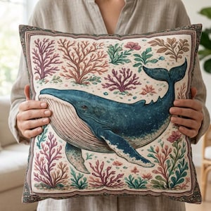 May include: Square decorative pillow featuring a blue whale illustration surrounded by coral and seaweed in shades of red, green, and brown. The pillow has a cream-colored background and a patterned border. A nautical-themed home decor item.