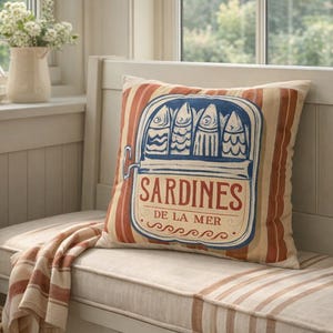 May include: Cushion with a sardine can design. The pillow has a cream background with red and tan vertical stripes. The sardine can graphic is blue and cream with the word "Sardines" in red. The text "De La Mer" is also included.