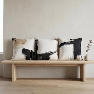May include: Three square cushions with abstract black and white designs are arranged on a light wood bench. One cushion has tan accents. A small vase with greenery sits on the bench.