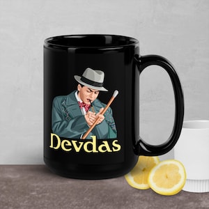 May include: Black ceramic mug with a white image of a man in a hat smoking a cigarette and holding a cane. The text "Devdas" is printed in yellow below the image.