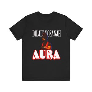 May include: Black t-shirt featuring the text "DILJIT DOSANJH" above a graphic of a person wearing a black head covering and a red vest. The word "AURA" is printed in large, red, outlined letters below the graphic.