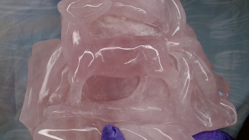 May include: A pink rose quartz sculpture of a cow, carved in a layered style. The sculpture is polished and translucent, with light shining through the stone.