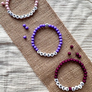 Könnte beinhalten: Drei Perlenarmbänder mit verschiedenen Perlenfarben und weißen Buchstabenperlen, die "SCHNEEHAASE", "CHEERS" und "APRES CHIC" buchstabieren.