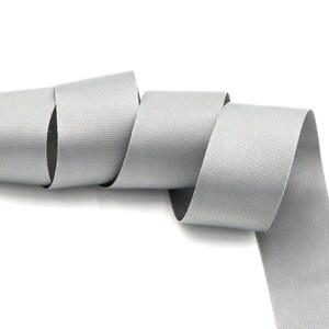 May include: A coiled length of light gray webbing material, likely nylon or a similar synthetic. The webbing has a textured surface with a subtle dotted pattern. The material is rolled up on a white background.