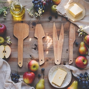 May include: A selection of wooden serving utensils, including a spatula, server, and knife, are displayed on a rustic wooden surface. The image is enhanced by fresh fruit, cheese, and olive oil, creating a natural and inviting aesthetic.