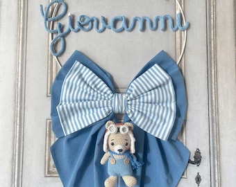 May include: A blue and white striped bow hangs from a white metal wreath with the name "Giovanni" spelled out in blue yarn. A small crocheted teddy bear wearing a pilot's hat and overalls is attached to the bow. The wreath also has a blue cloud with the text "è nato" and a small orange and gray airplane.