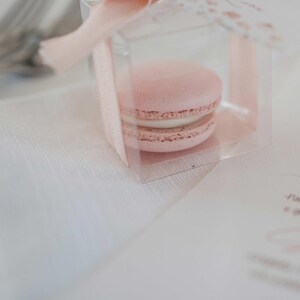 May include: A pink macaron in a clear plastic box tied with a pink ribbon. The box is sitting on a white table with a menu card.