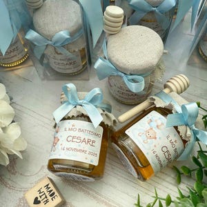 Peut inclure: Petits pots de miel avec des couvercles en toile de jute et des rubans bleu clair. Les étiquettes indiquent "IL MIO BATTESIMO CESARE 16 NOVEMBRE 2025". Chaque pot contient un bâtonnet à miel en bois et est présenté dans une boîte transparente.