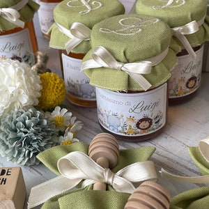 May include: Green fabric favour bags with white ribbon and wooden honey dippers. The bags are filled with small jars of honey with green fabric lids and labels that say "Il battesimo di Luigi".