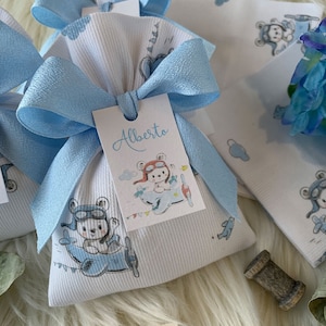 May include: A white fabric gift bag with a blue ribbon and a tag that says "Alberto". The bag has a pattern of bears flying aeroplanes. The bag is sitting on a white fluffy surface. A small brown spool of thread is in the background.