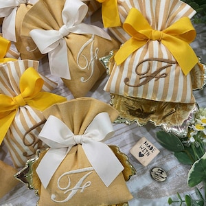 May include: A collection of small, yellow fabric bags, each with a white embroidered initial and a white bow. Some bags feature yellow and white striped fabric. The bags are presented on gold-coloured leaf-shaped trays.