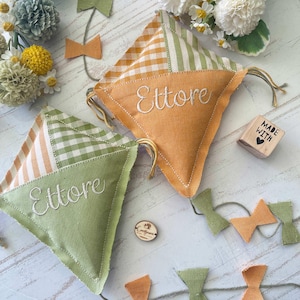 May include: Two green and orange fabric kites with the name Ettore embroidered on them. The kites are surrounded by a garland of green and orange fabric bows. There is a small wooden block with the words "Made with" on it.