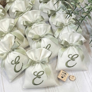 May include: Off-white fabric bags, each tied with a sage green satin ribbon and embroidered with a green cursive letter "E". The bags are arranged with eucalyptus and a wooden stamp that says "MADE WITH" and a heart.