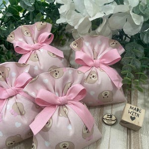 May include: Four pink fabric gift bags with a brown teddy bear print and pink ribbon bows. The bags are sitting on a wooden surface with a small wooden block that says "Made with ♥".