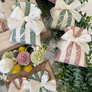 May include: A collection of striped fabric bags in shades of green and pink, adorned with off-white bows. Each bag is embroidered with the letter "L". The bags are presented with floral arrangements, suitable for gifting.