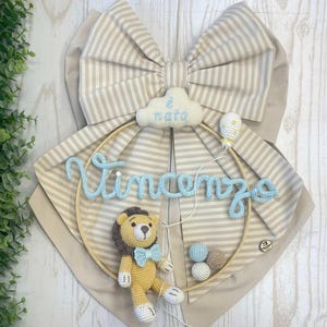 May include: A handmade nursery decoration featuring a crocheted lion with a blue bow tie, the name "Vincenzo" in light blue script, and a cloud with the text "è nato". The design includes a striped bow and a wooden hoop.