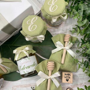 May include: A collection of small jars and bags, possibly favours, in a light green colour scheme. The jars have green fabric lids embroidered with a white "E" and tied with white ribbon. Small wooden honey dippers are included.