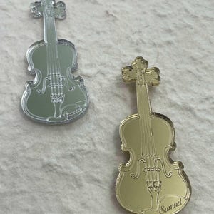 Könnte beinhalten: Zwei spiegelnde Violin-Anhänger, einer silber und einer gold. Die silberne Violine hat den Namen "Gracelyn" eingraviert. Die goldene Violine hat den Namen "Samuel" eingraviert.