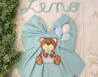 May include: A light blue fabric bow with a brown teddy bear holding balloons. The bow is attached to a white metal hoop with the name "Zeno" written in white yarn. The hoop is decorated with a blue and white striped cloud and white stars.
