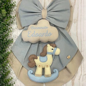 May include: A baby shower decoration featuring a light blue bow with a beige accent. A cloud-shaped ornament with the name "Edoardo" embroidered in blue hangs below the bow. A rocking horse is attached to the decoration.