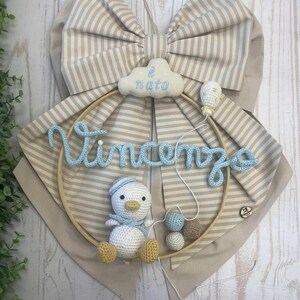 May include: A handmade nursery decoration featuring a large striped bow in beige and white. A wooden hoop holds a crocheted duck, the name "Vincenzo" in blue yarn, and the text "è nato" on a cloud.